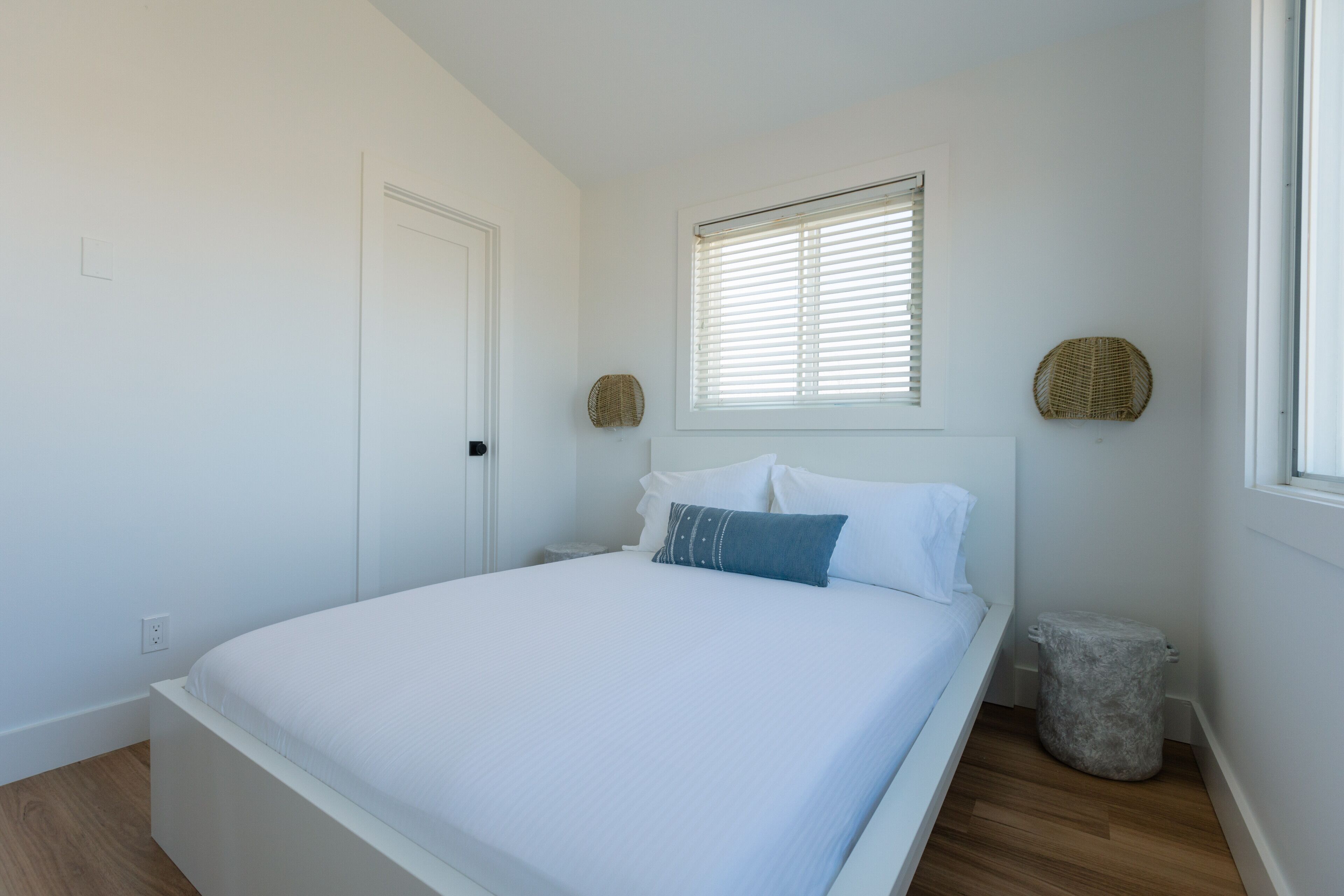 A bedroom with a bed, a window with blinds, a door, and a small round wicker basket on the wall.