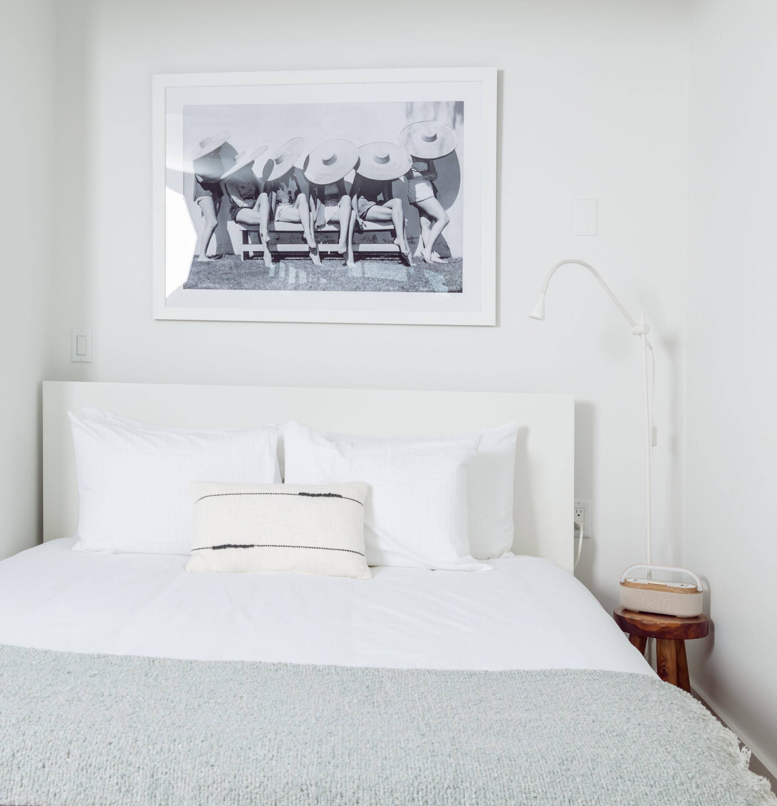 A neatly made bed with white linens and a single pillow, a bedside table with a lamp, and a framed black and white photograph above the bed.