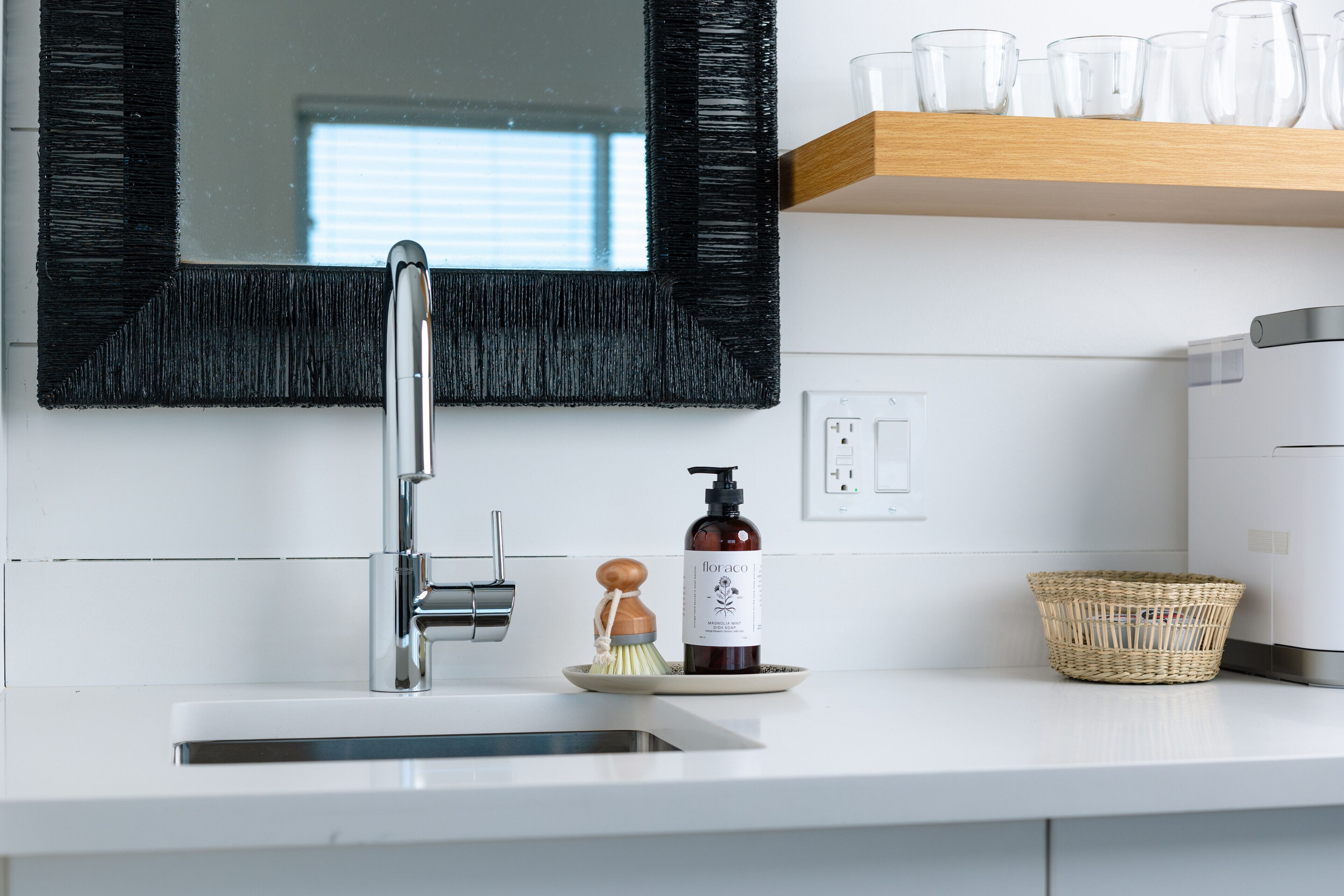 A modern kitchen with a stainless steel sink, a black-framed mirror, and a white countertop.