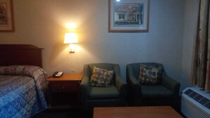 A hotel room with a bed, a nightstand with a lamp, two armchairs with patterned cushions, a small table, and a wall-mounted air conditioner.