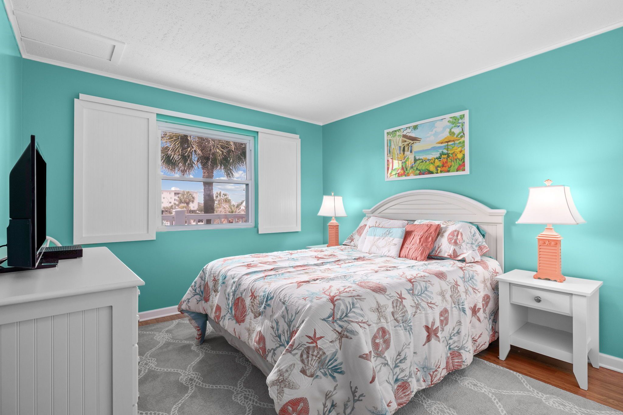 A bedroom with teal walls, a bed with floral bedding, two bedside lamps, a window with a view, and a dresser with a TV.