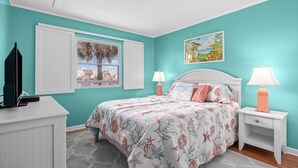 Une chambre avec des murs de couleur turquoise, un lit avec une literie à motifs floraux, deux lampes de chevet, une fenêtre donnant sur un beau paysage et une commode sur laquelle est posée une télévision.