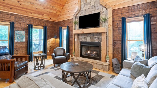 A cozy cabin interior with a stone fireplace, wooden walls, leather furniture, and a large window.