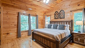 A wooden cabin bedroom with a bed, a ceiling fan, and two windows with curtains.