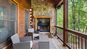 A wooden deck with wicker furniture, a stone fireplace, and a view of the forest.