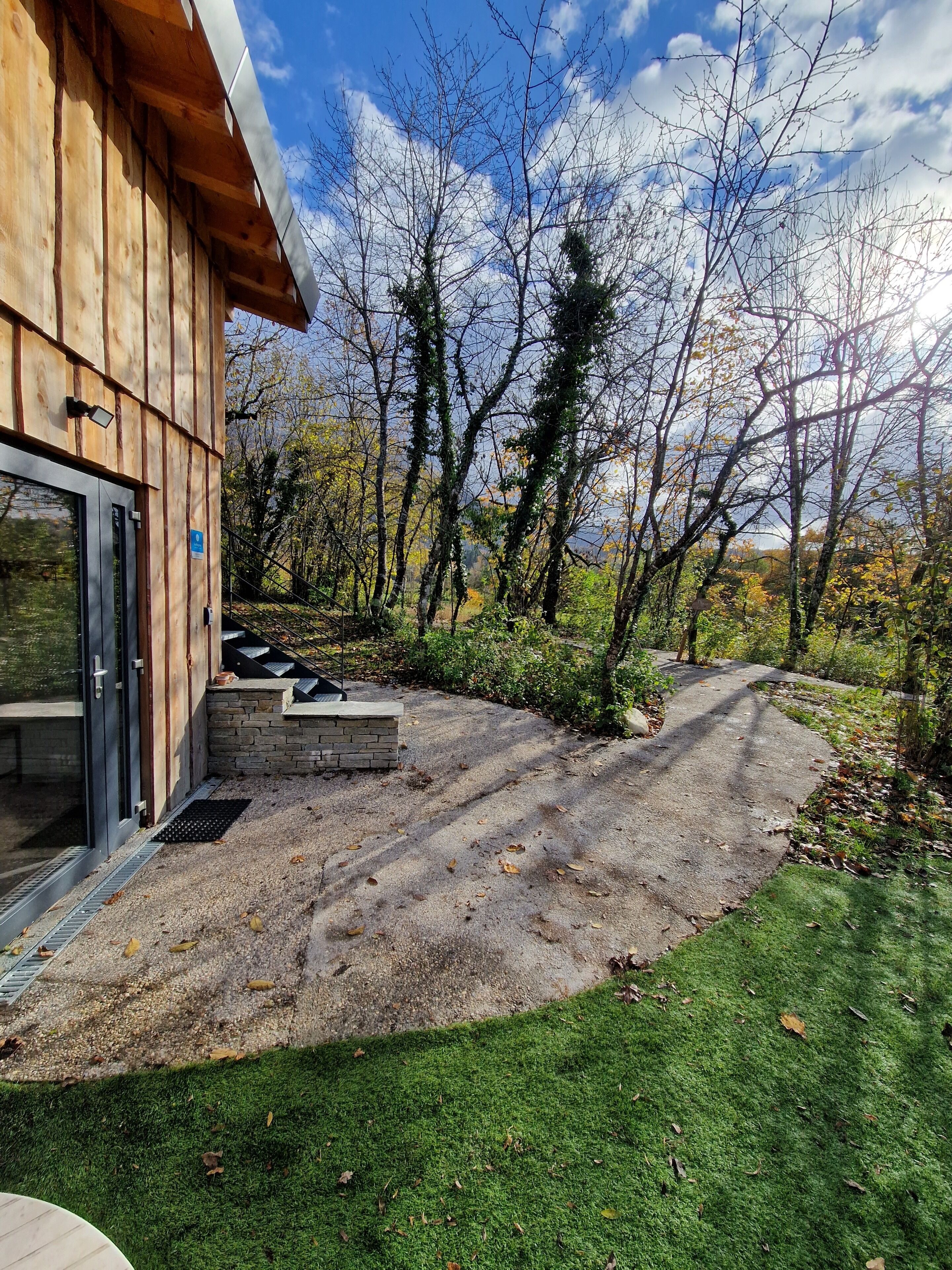 Un bâtiment en bois avec une entrée dotée d’une porte en verre, un chemin de pierre menant à une zone herbeuse, et, au fond, une vue sur des arbres sans feuilles.