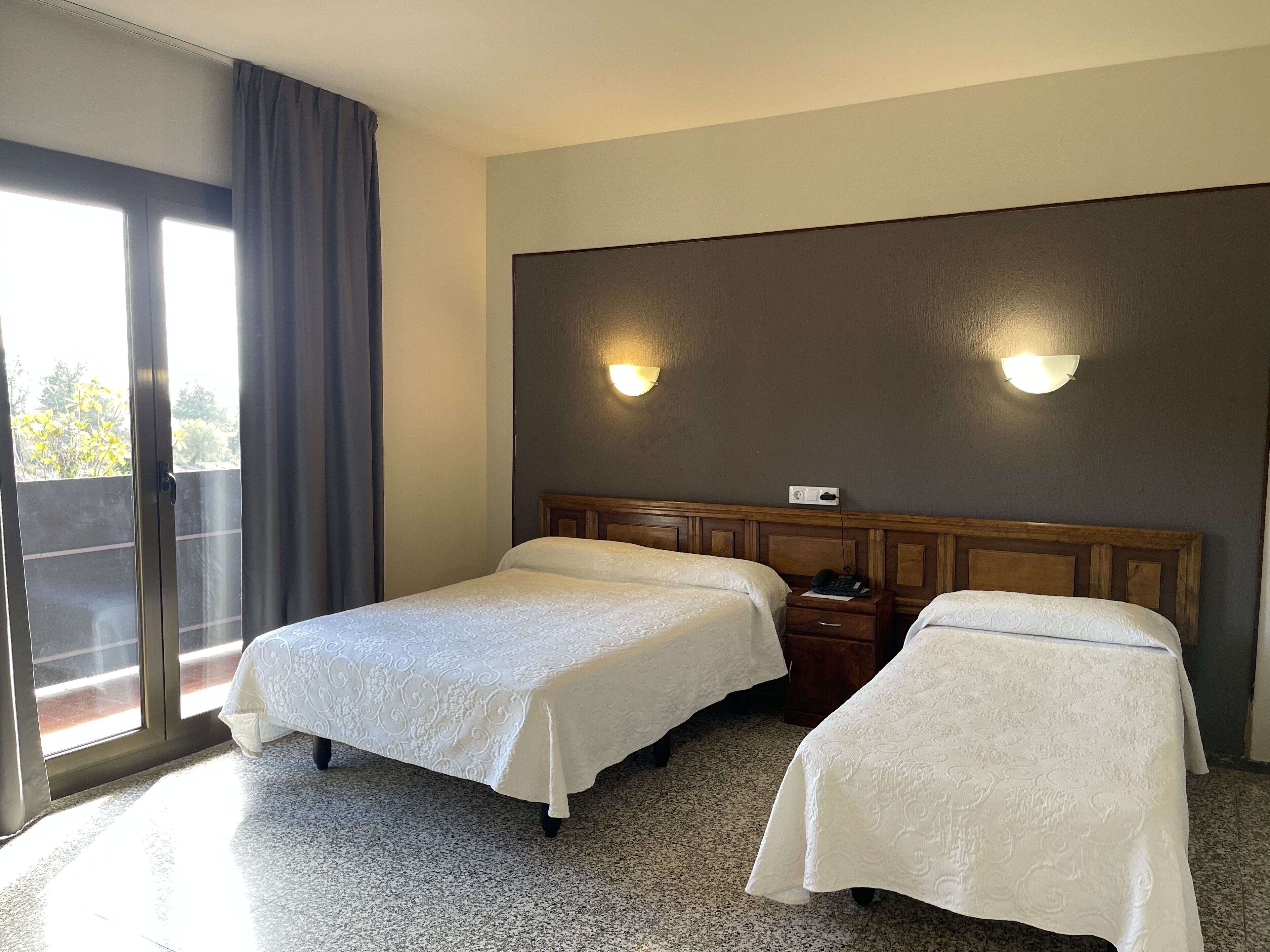 Una habitación de hotel con dos camas, un ventanal con cortinas y una lámpara fijada a la pared.