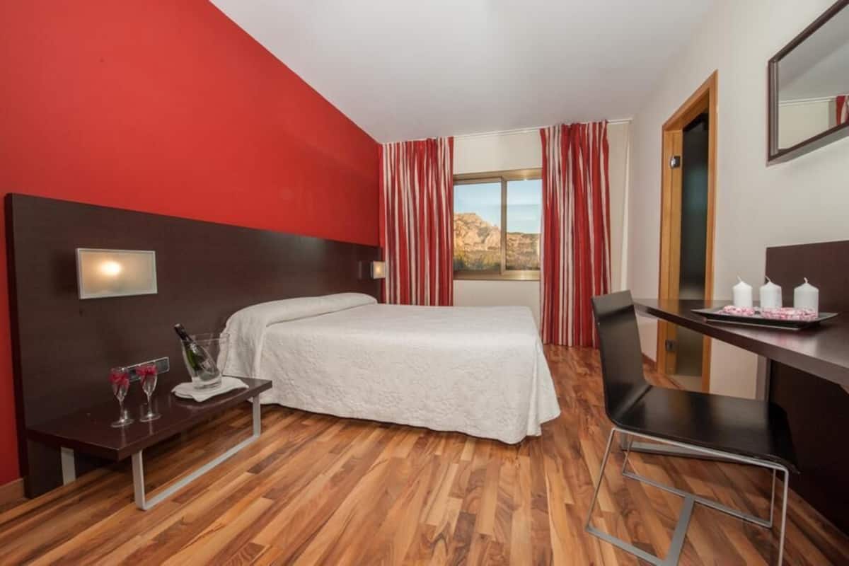 Quarto de hotel com cama, escrivaninha, cadeira, mesinha com garrafa e copos, e janela com cortinas.