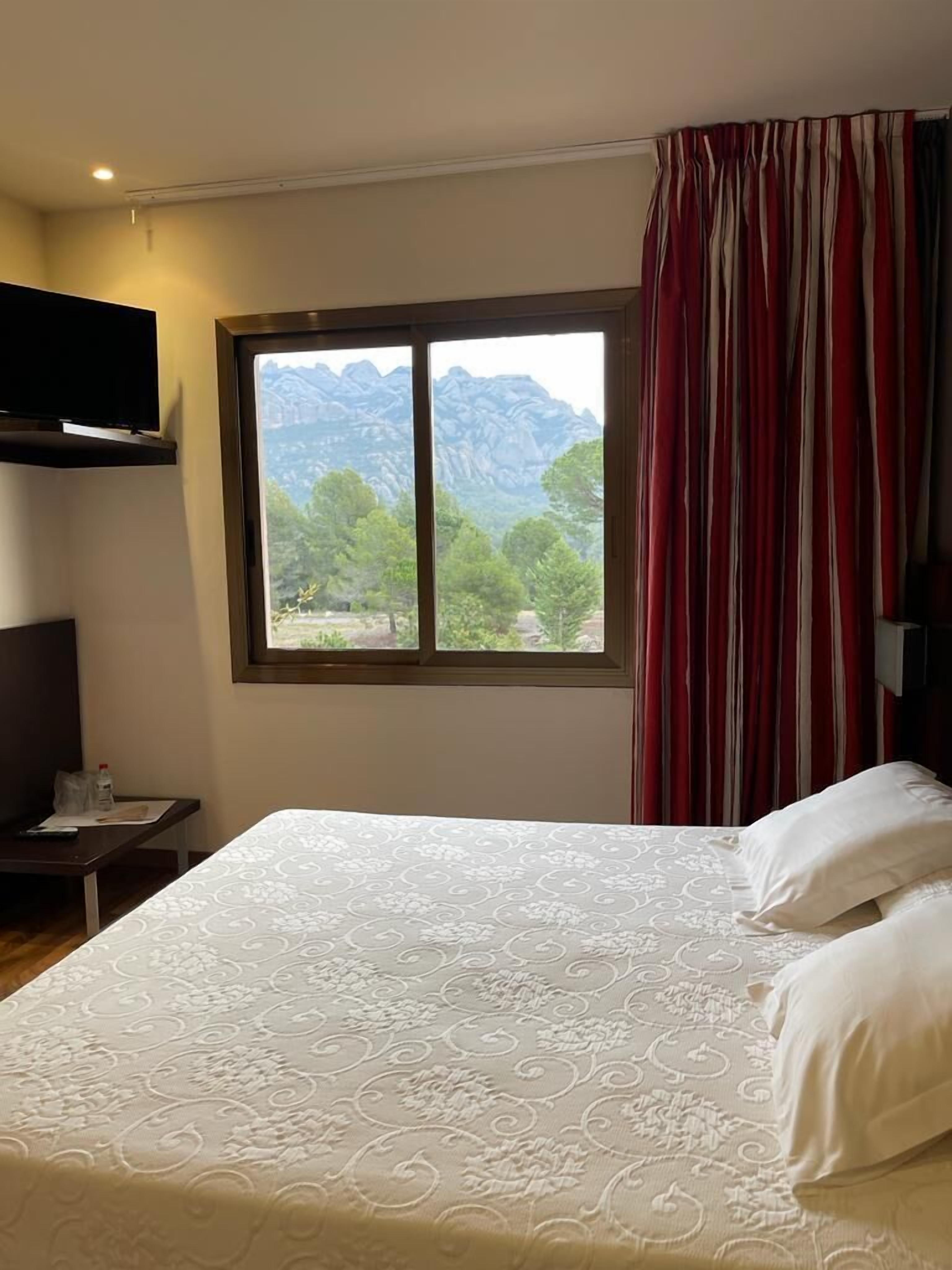 A hotel room with a large bed, a TV mounted on the wall, and a window with a mountain view.