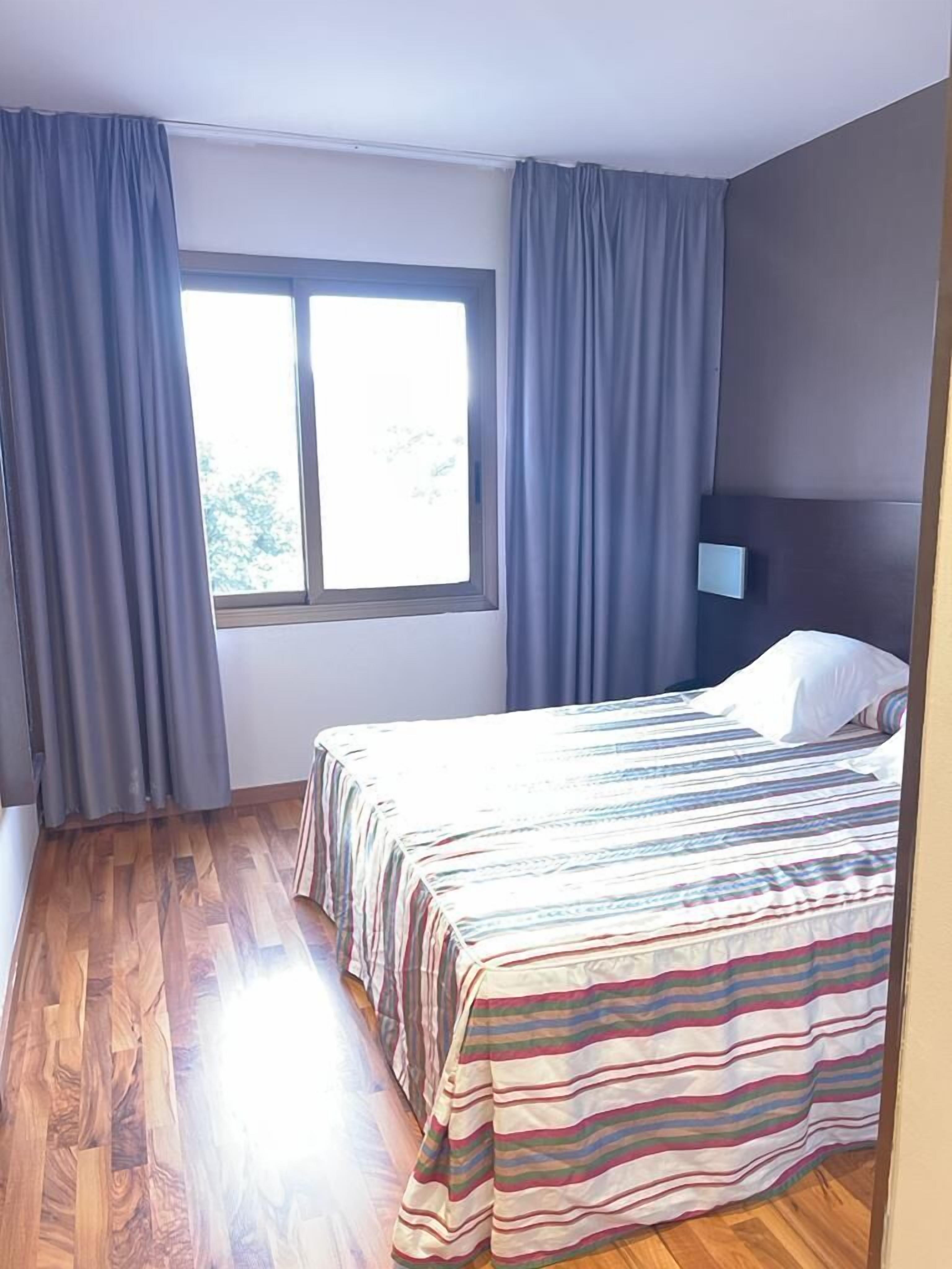 Une chambre à coucher avec un lit, des rideaux, une fenêtre et une lampe fixée au mur.