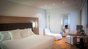 Una habitación de hotel moderna con una cama grande, un área de descanso con una silla y una mesita, y un espejo en la pared.