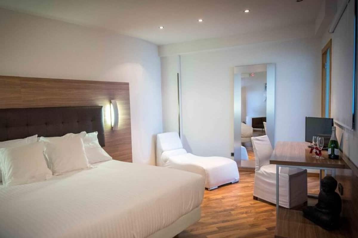 Quarto de hotel moderno com uma cama grande, uma área de estar com uma poltrona e uma mesa pequena, e um espelho na parede.