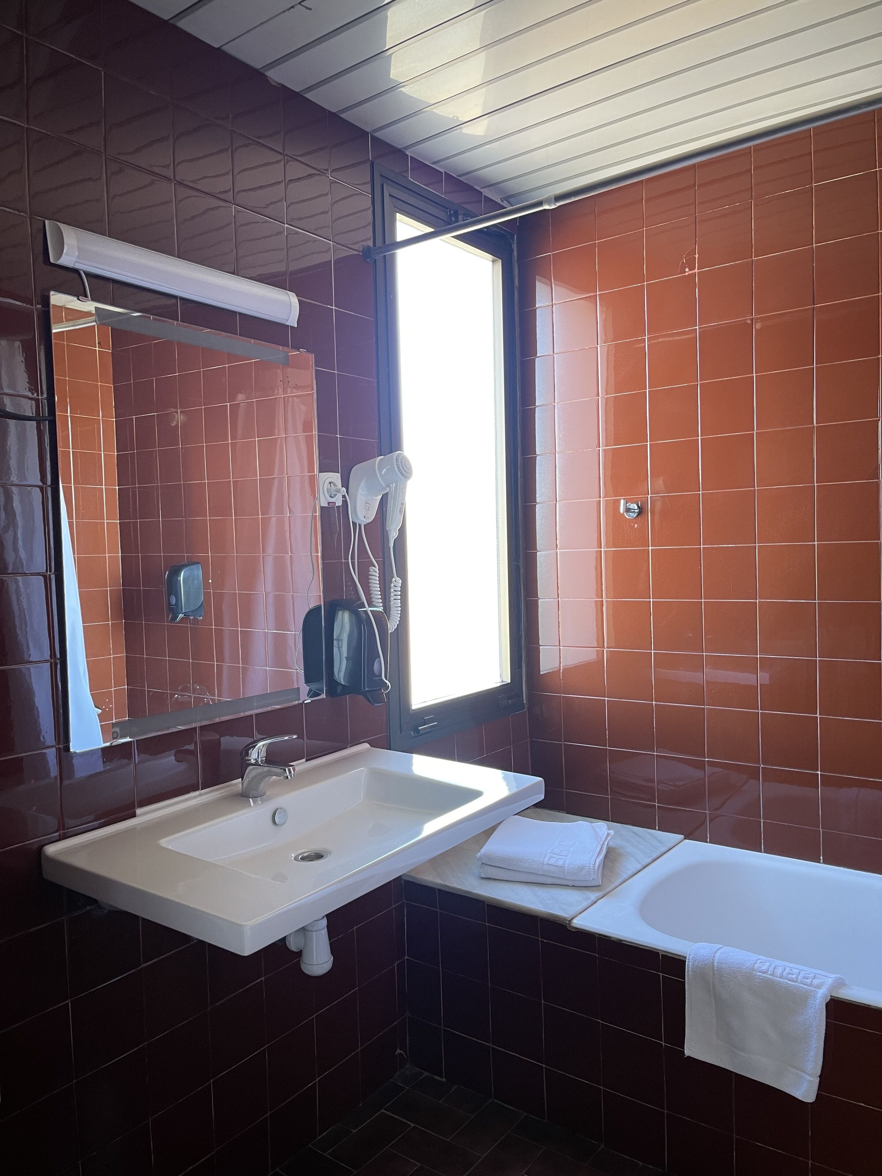 Une salle de bain avec un lavabo blanc, un miroir, une serviette et une fenêtre.