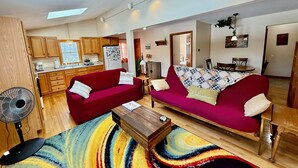 Un salón espacioso con un sofá rojo, una mesa de centro de madera y una alfombra colorida. Se aprecia una cocina al fondo, con armarios de madera y un refrigerador.