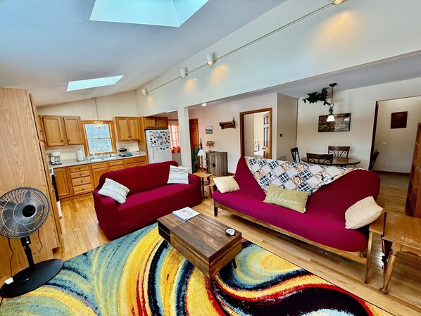 A spacious living area with a red sofa, a wooden coffee table, and a colorful rug. There is a kitchen in the background with wooden cabinets and a refrigerator.