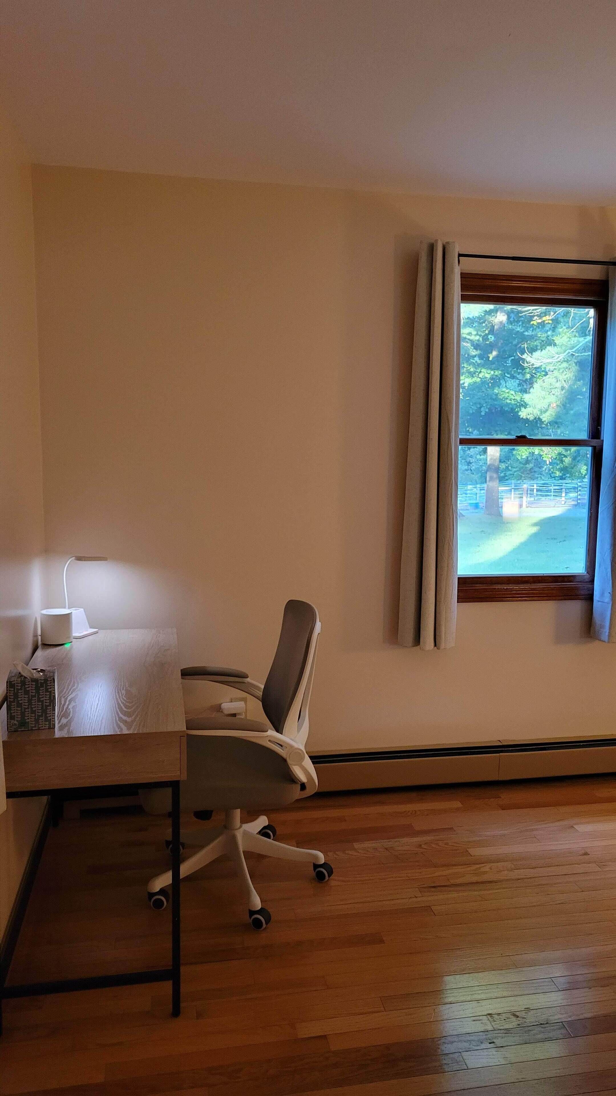A room with a wooden floor, a desk with a lamp, a chair, and a window with curtains.