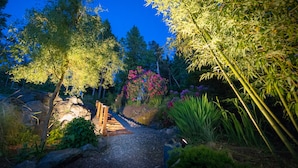 A garden path with a wooden bench, surrounded by various plants and trees, leading to a rocky area.