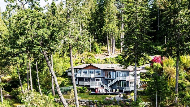 A two-story house with a balcony, surrounded by dense forest and a rocky shoreline.
