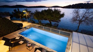 Una piscina rectangular con fondo de cristal, rodeada de sillones de descanso y una pequeña mesa, con vistas a una masa de agua, árboles y colinas al fondo.
