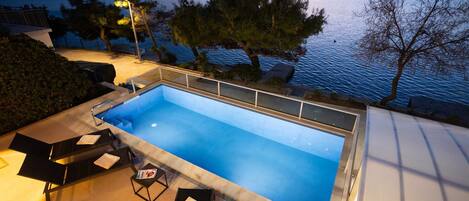 A rectangular swimming pool with a glass bottom, surrounded by lounge chairs and a small table, overlooking a body of water with trees and hills in the background.