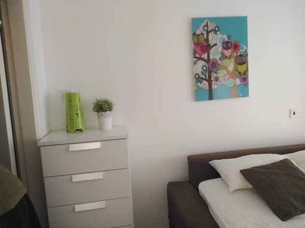 Un dormitorio con una cama, un aparador, una pequeña planta en maceta y un cuadro colgado en la pared que muestra pájaros coloridos en un árbol.
