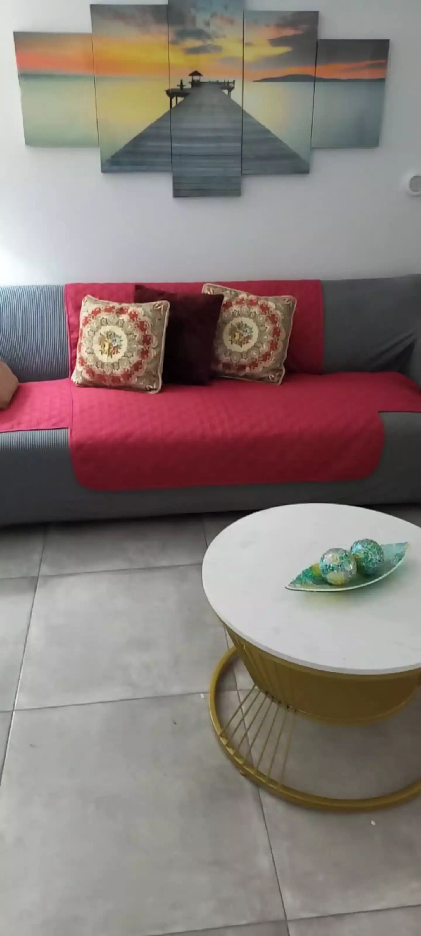 A living room with a grey sofa, red throw, patterned cushions, a round coffee table with decorative items, and a sunset painting above.