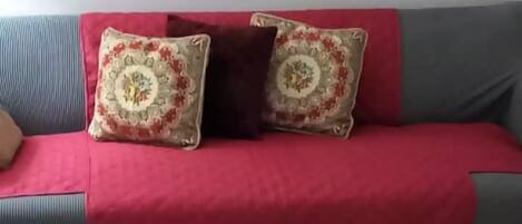 A living room with a grey sofa, red throw, patterned cushions, a round coffee table with decorative items, and a sunset painting above.