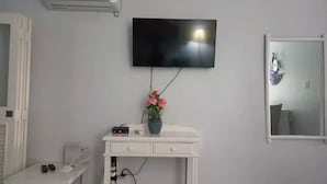 A room with a white desk, a flat-screen TV mounted on the wall, a vase with flowers, and an air conditioning unit.