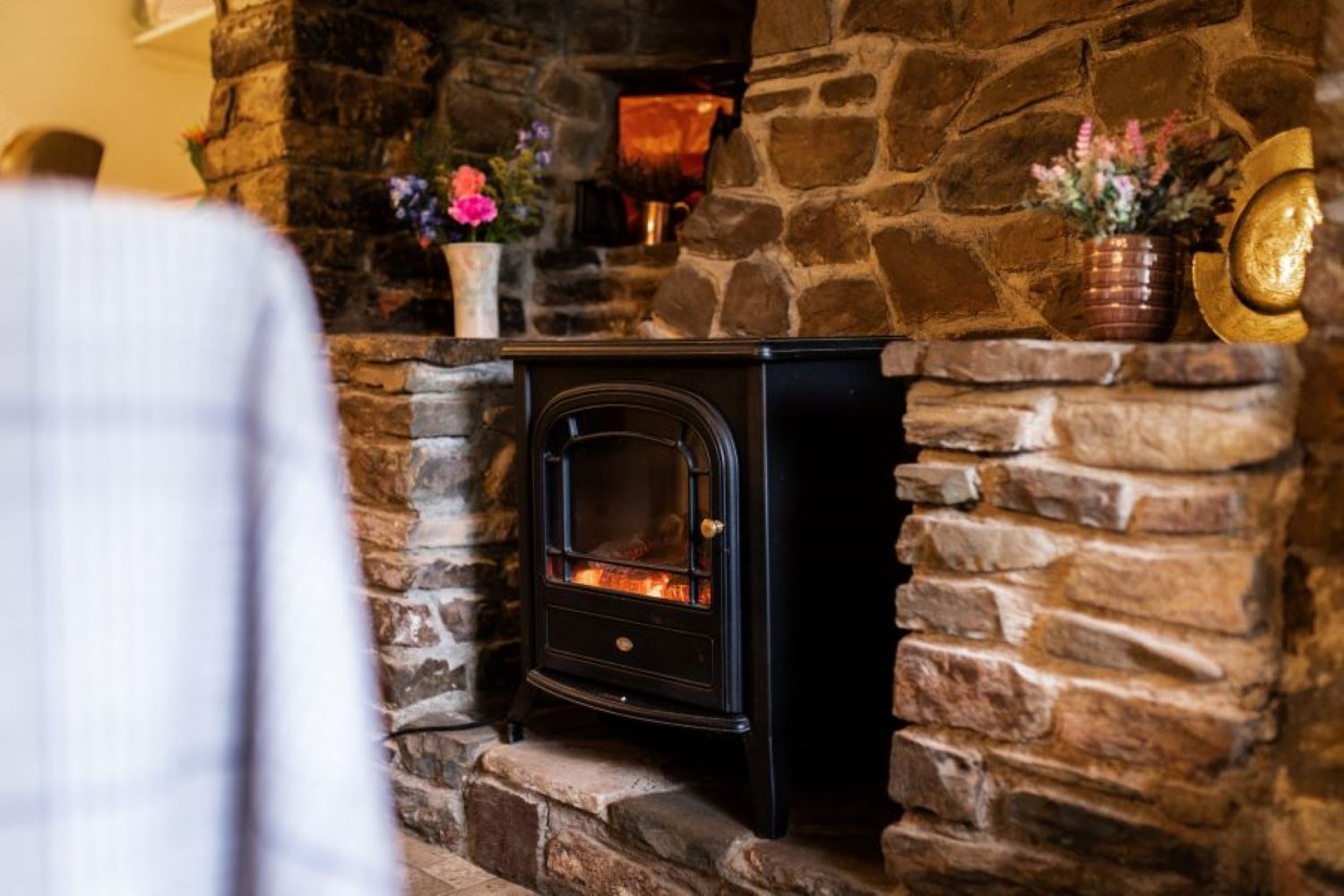 A stone fireplace with a black wood-burning stove, surrounded by stone walls and decorated with flowers.