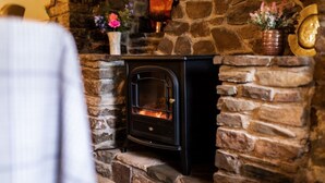 A stone fireplace with a black wood-burning stove, surrounded by stone walls and decorated with flowers. - 2 bed rural retreat nestled in the heart of Exmoor (Parracombe)