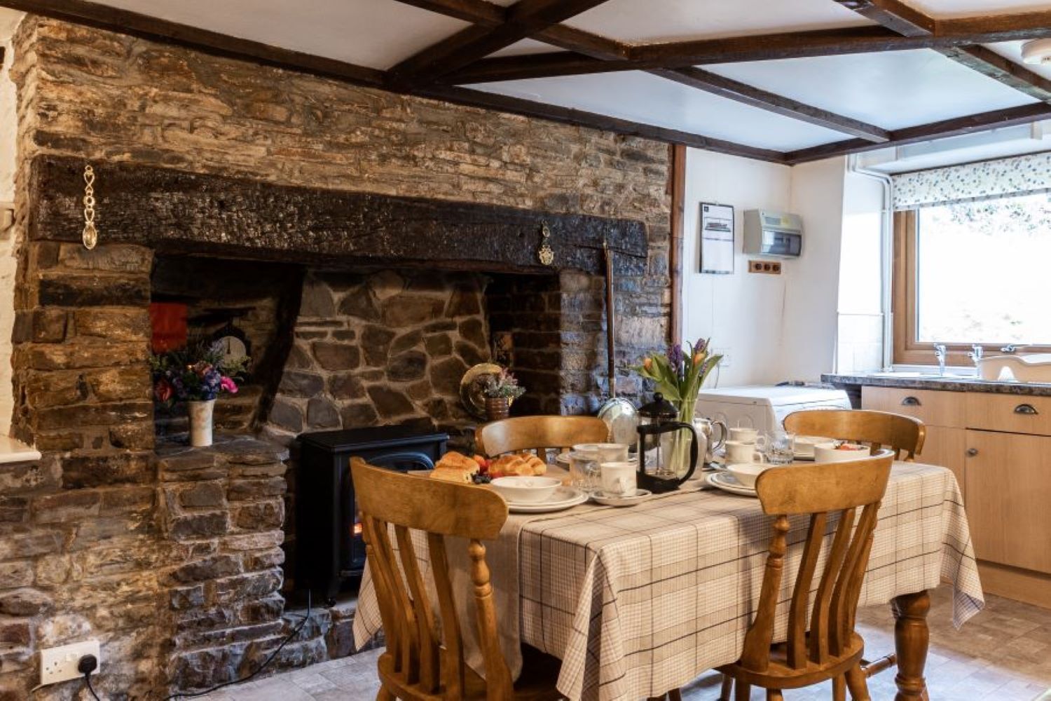 Un rincón comedor acogedor con chimenea de piedra, sillas de madera, una mesa dispuesta para el desayuno y una cocina al fondo.