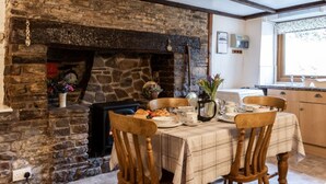 A cozy dining area with a stone fireplace, wooden chairs, a table set for breakfast, and a kitchen in the background. - 2 bed rural retreat nestled in the heart of Exmoor (Parracombe)