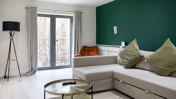 Un salon moderne avec un canapé d’angle, une petite table basse ronde, un lampadaire et une vue à travers une porte vitrée.