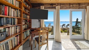 Une pièce avec une bibliothèque en bois, un téléviseur, un bureau en bois et une chaise. On peut admirer la mer à travers une porte vitrée.