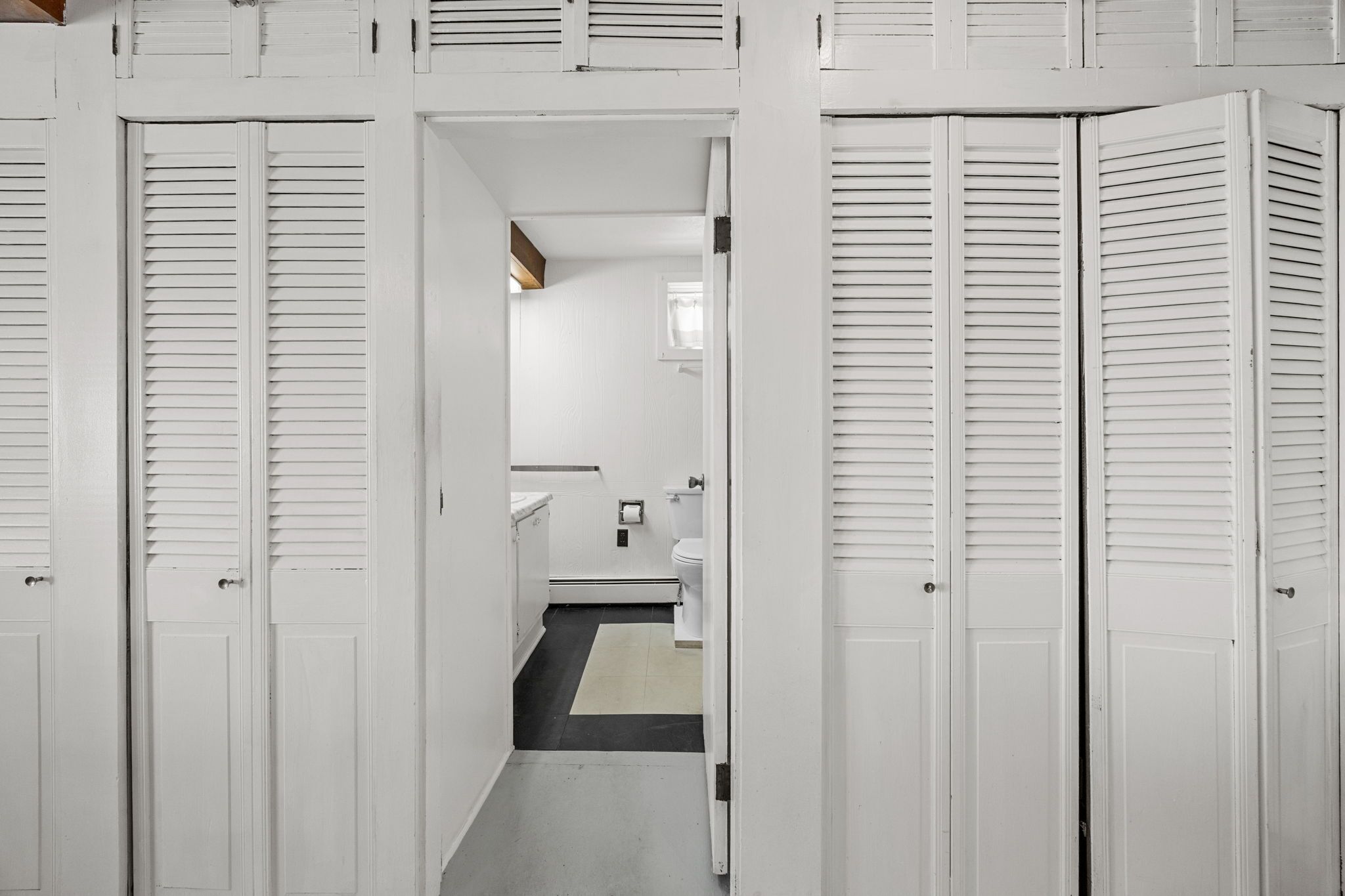 A narrow hallway with white doors and shutters, leading to a bathroom with a toilet, sink, and towel rack.