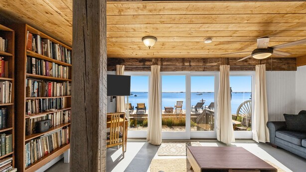 Un salon chaleureux avec un plafond en bois, une bibliothèque, un téléviseur à écran plat, un canapé, une table basse et une vue sur la mer grâce à de grandes portes vitrées.