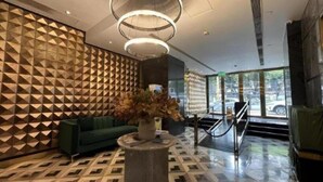 Eine moderne Lobby mit einer Wand im geometrischen Muster, einem grünen Sofa, einem runden Holztisch mit einer Vase und einer Treppe, die zu einer oberen Ebene führt.