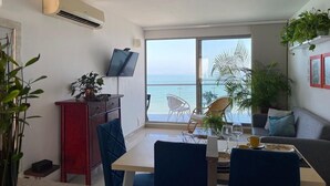 Un soggiorno moderno con zona pranzo, ampie finestre vista mare, televisore piatto a parete e diverse piante in vaso.
