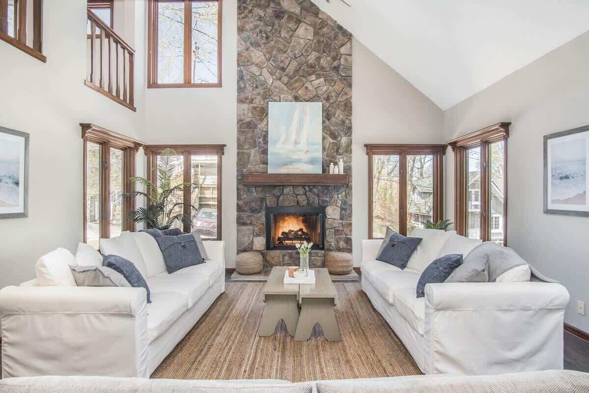 Una sala de estar con dos sofás blancos, una chimenea de piedra y ventanas amplias.