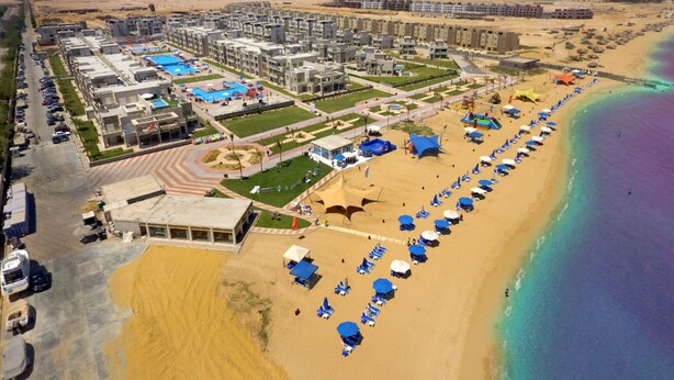 Vista aérea de un resort costero con varias edificaciones, una playa de arena con sombrillas y una piscina.