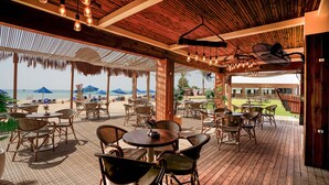 Un patio al aire libre con mesas y sillas de madera, techo de paja y vista a la playa.