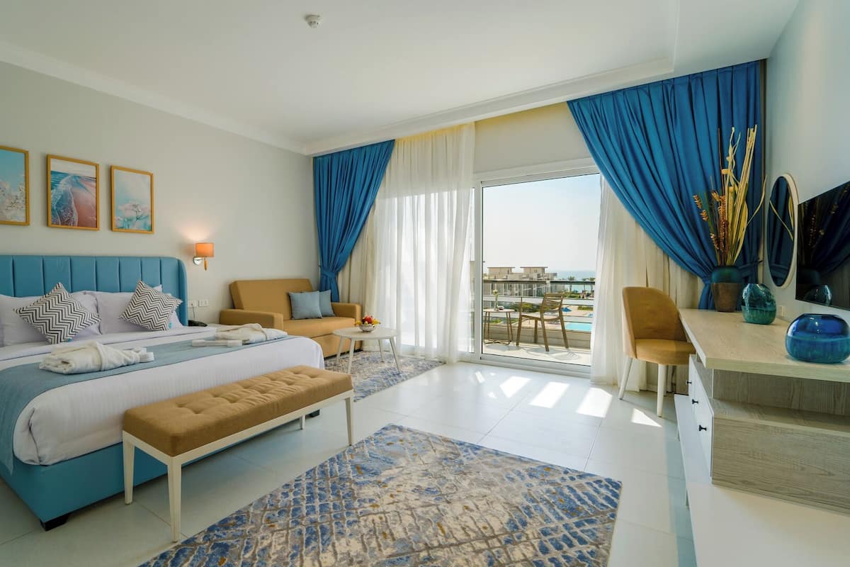 Ein modernes Hotelzimmer mit einem großen Bett, einer Couch, einem Schreibtisch und einem Balkon mit Blick auf den Pool.