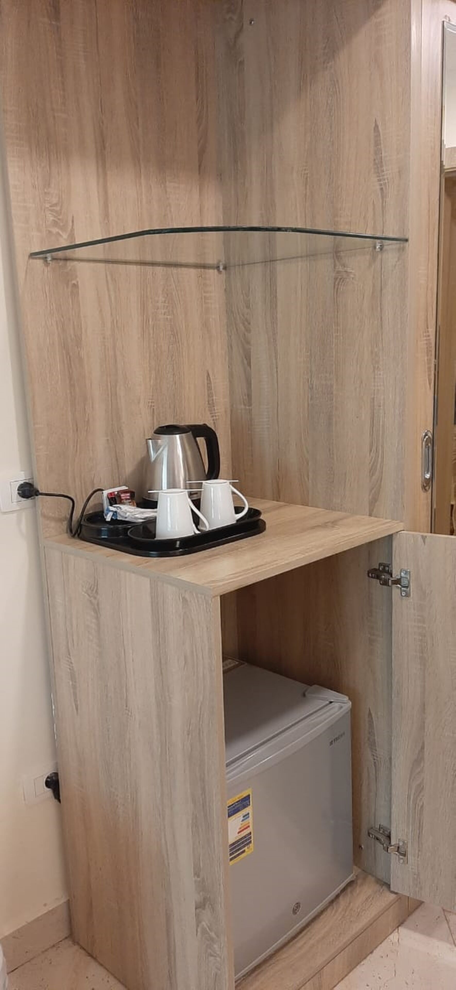 A compact kitchenette with a wooden cabinet, a small refrigerator, and a glass shelf holding a kettle and cups.