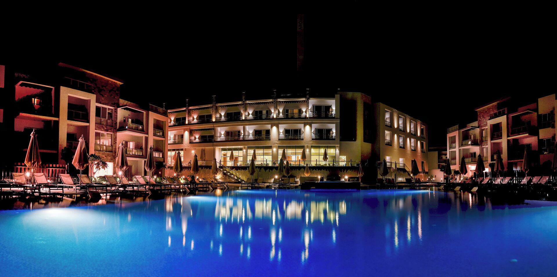 A nighttime view of a hotel with a swimming pool, multiple balconies, and outdoor seating areas.