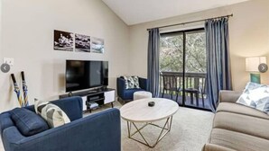 Een woonkamer met een televisie, blauwe banken, een ronde salontafel en een balkon met gordijnen.