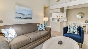 Een woonkamer met een beige bank, een blauwe fauteuil, een ronde salontafel en een plafondventilator.