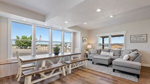 Un salon spacieux avec une table et des bancs en bois, un canapé gris et de grandes fenêtres donnant sur un espace extérieur clos.