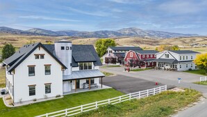 A white two-story house with a covered porch, a red barn, and a view of mountains in the background. - Cedar Farmhouse and Silo w/POOL (Sleeps 50) (Garden City)