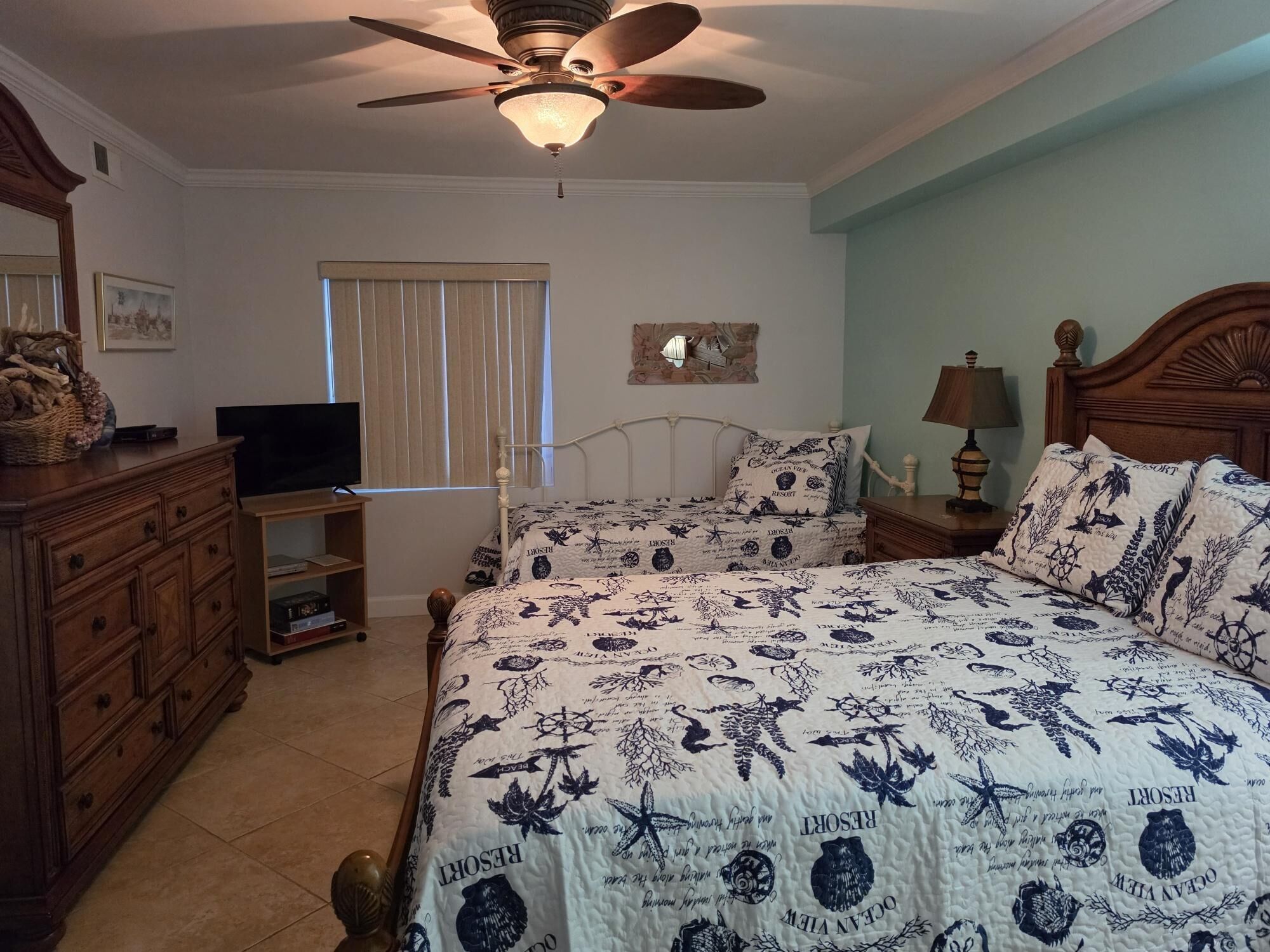 Une chambre à coucher comprenant un lit, une commode, un téléviseur, un ventilateur de plafond et une fenêtre avec des stores.