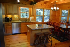 A wooden cabin kitchen with granite countertops, a dishwasher, a coffee maker, and a dining area with red chairs and a table. - Riverfront cabin with WiFi, 2 fireplaces, game room, pool table and hot tub. (Ellijay)